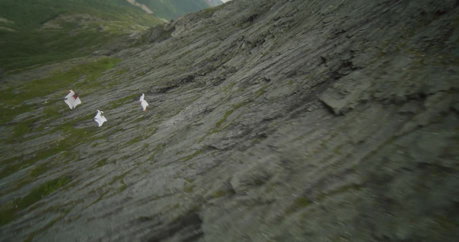 Three people wearing wingsuits are floating along the side of a mountain toward a lake in the valley