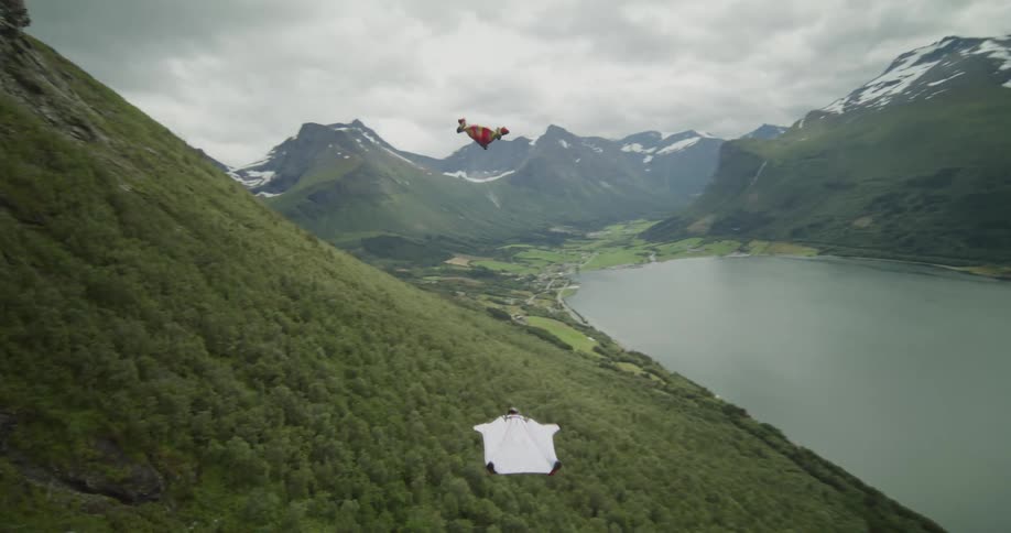 Two people wearing wingsuits are floating along the side of a mountain toward a lake in the valley