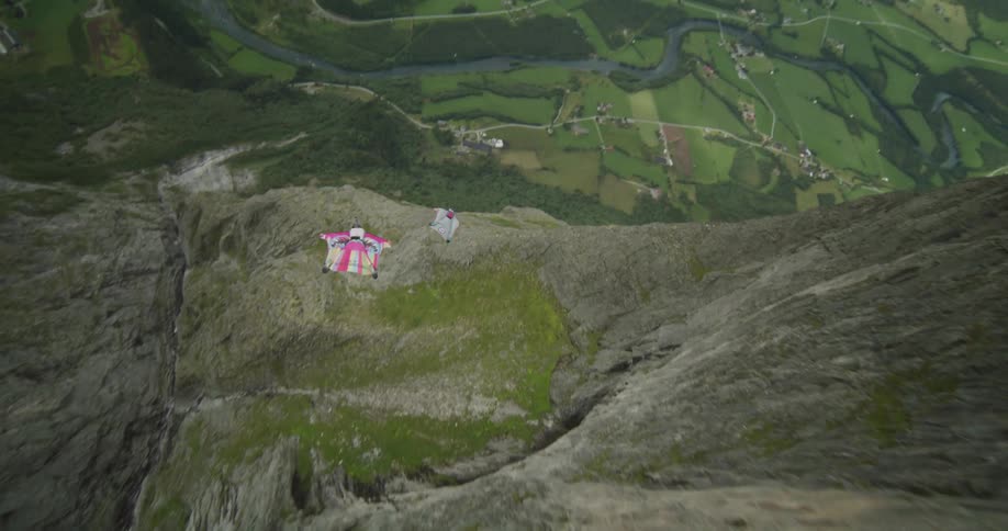 Two people wearing wingsuits jump off a cliff and float along the side of the cliff and over the valley below