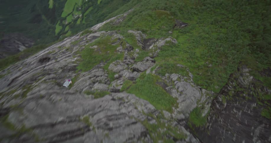 A person wearing a wingsuit is floating along the side of a mountain as they fly toward a mountain river valley
