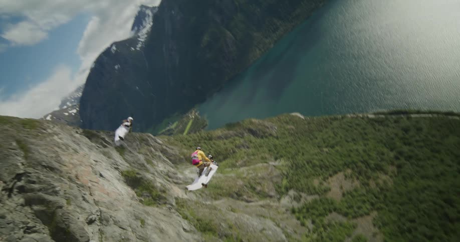 Person wearing parachute and people wearing wing suits glide and perform stunts over forest and lake near cliff