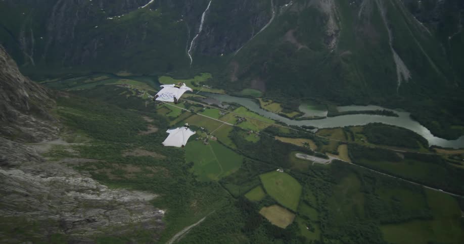 Two persons in white wingsuits gliding through the airover a valley and a river in a mountain region