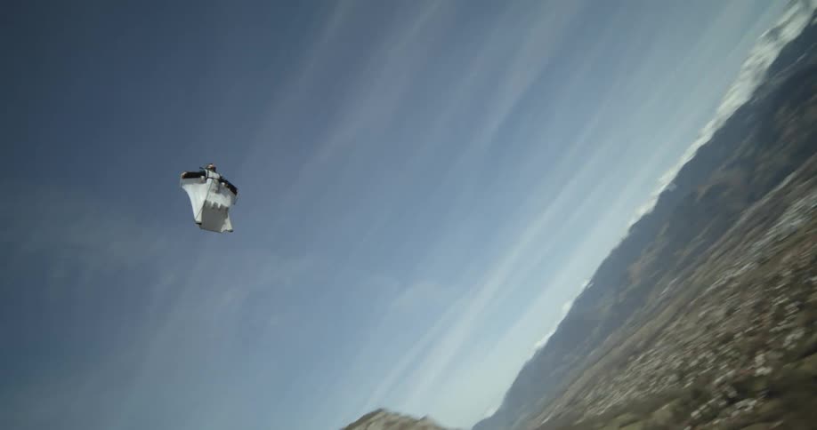A person wearing a wingsuit is flying along the side of a mountain as they open a parachute and float toward the valley community below