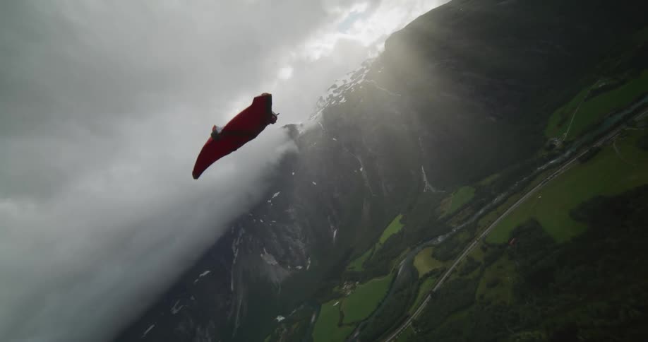 A person wearing a wingsuit is flying along the side of a mountain cliff as they float down to the valley below