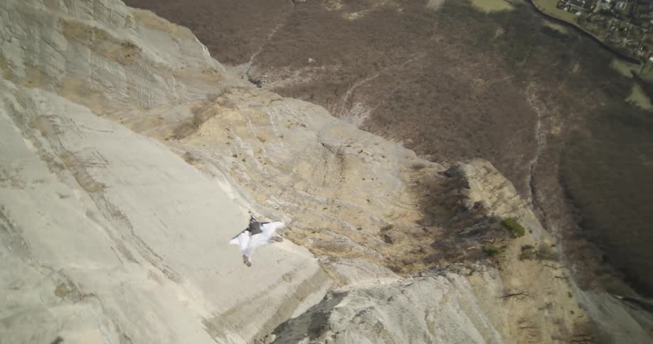 A person in a white suit jumps off a cliff spinning and then glides through the air