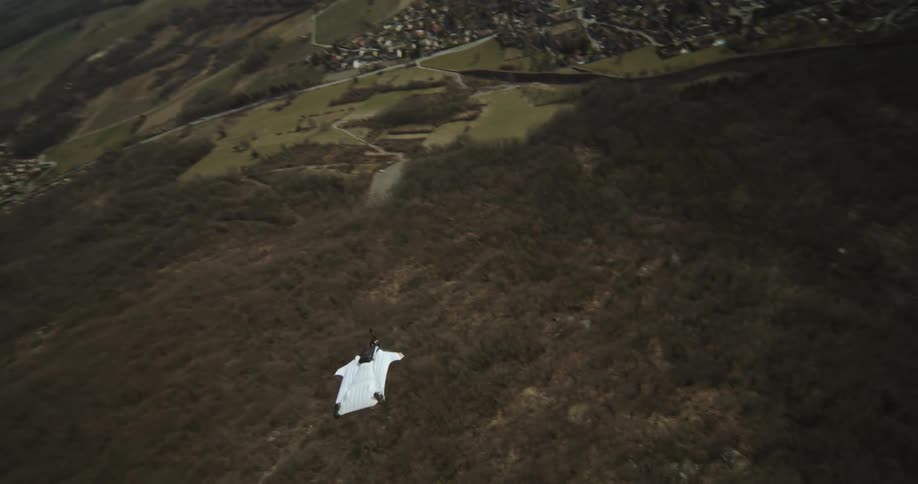 A person wearing a wingsuit jumps off a cliff and tumbles in the air and flips and then opens a parachute as they float toward the valley