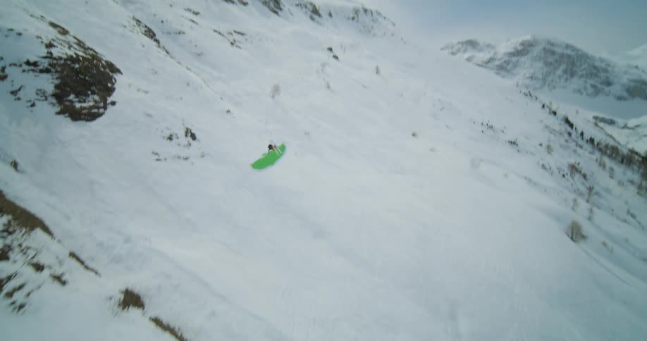 A person paraglides over a snowy mountain as two people ski down it