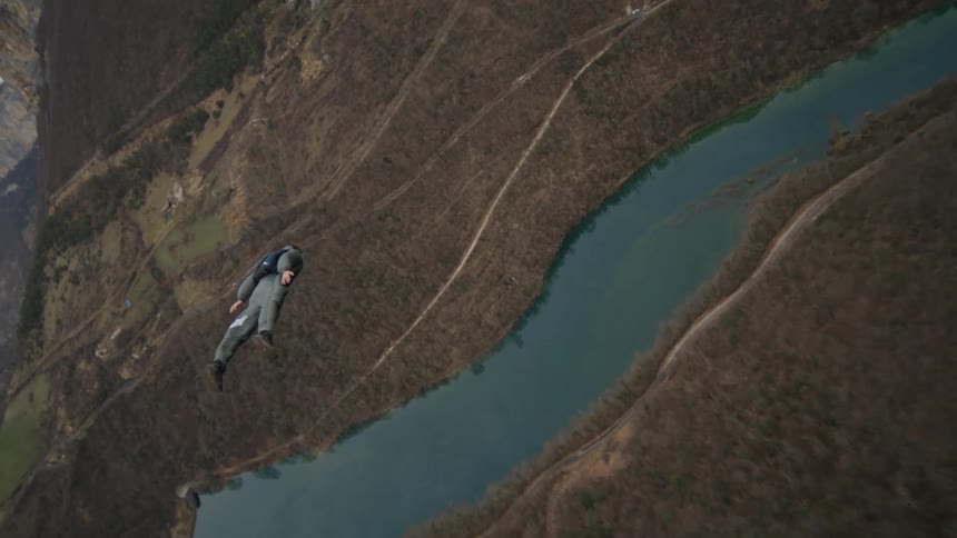 A person with a parachute pack on their back runs off the edge of a cliff and floats over the river valley below as they open their parachute