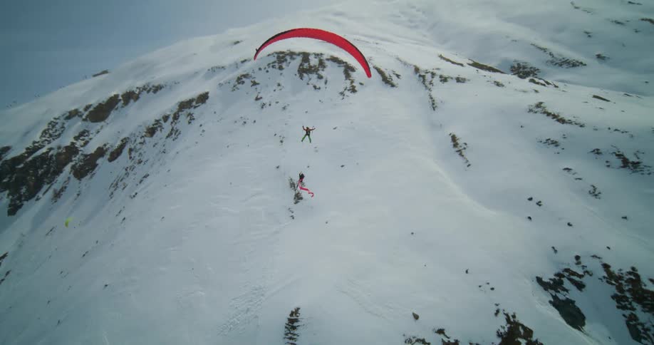 Two people hanging and gliding with a red parachute over snow covered mountains