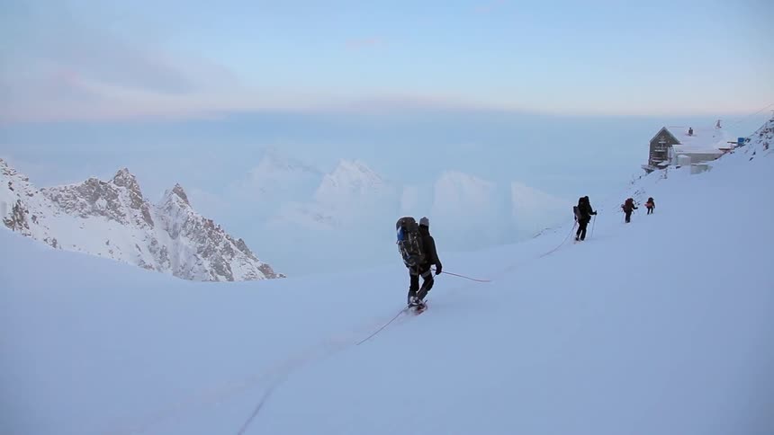 A group of mountain climbers ascend a snowy hill toward a building