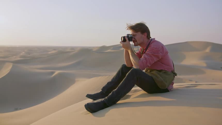 A photographer takes a picture while sitting on sand in a desert