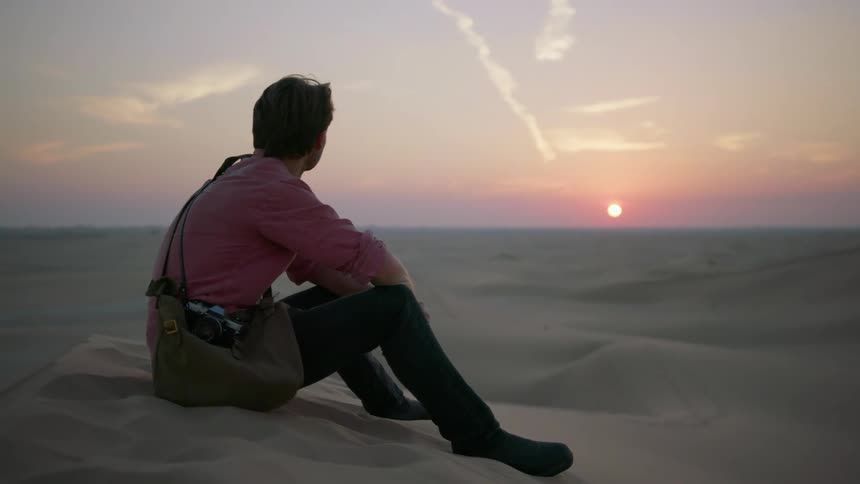 A man sits on a sand dune looking at the landscape at dusk