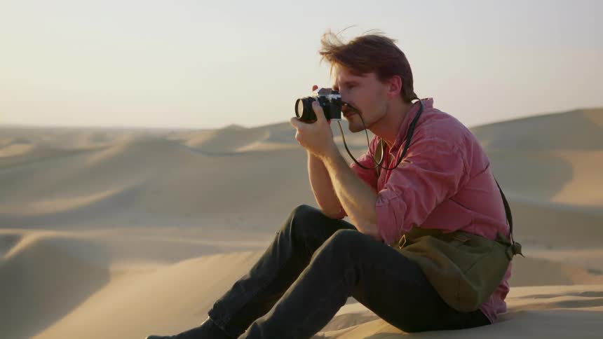 A man sitting on a sand dune takes a photo