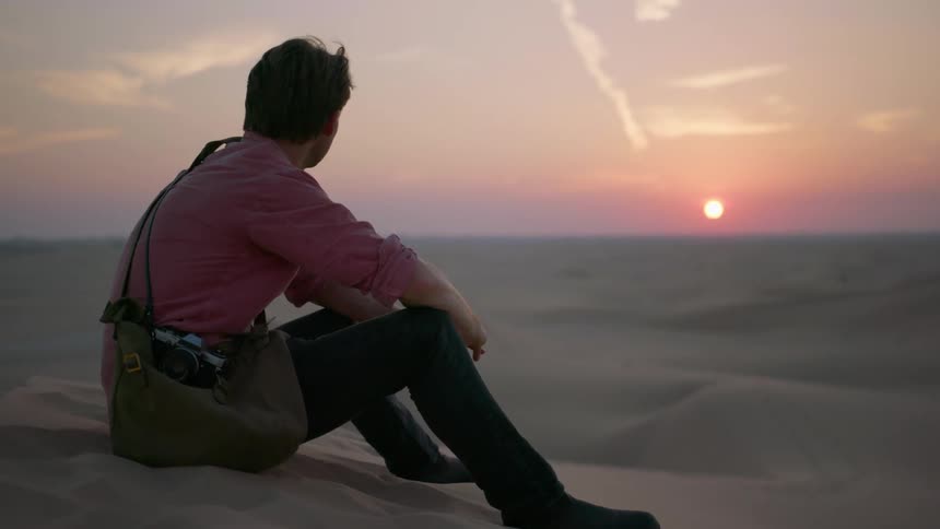 A man is sitting on the sand with a bag watching the sun set