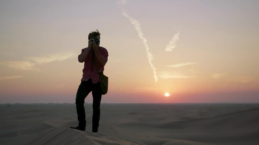Man standing on sand dunes taking photographs as sun sets