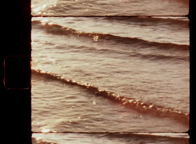 Super 8 close-up of small ocean or sea waves flowing and washing up or rolling onto the beach as the sun is shining with warm light and reflecting on the ripples of the water surface moving and rippling in a dynamic way