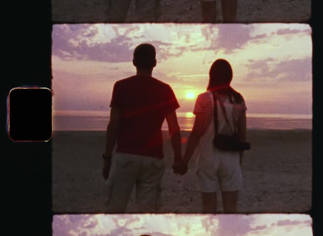 Man and woman holding hands standing on the beach near the sea watching sunset or sunrise on a romantic Valentine’s Day evening or morning date as the colorful sky glows red and purple