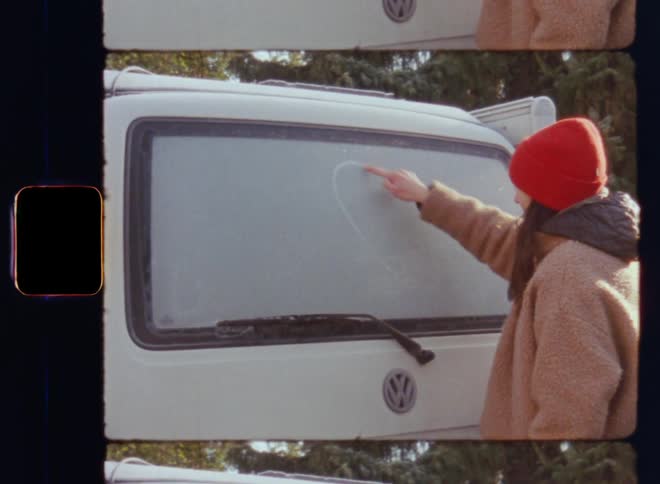 Romantic moment of a woman in love wearing a red woollen hat writing a heart with her hand and finger into the ice on the frozen rear window of a white T4 California Volkswagen or VW camper van on nostalgic Super 8mm film