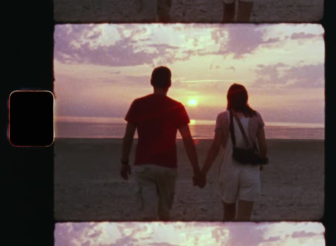 Romantic cliche moment of a love couple consisting of young man and woman in summer clothes holding hands while walking along a beach at sunset looking at the red and pink sky with clouds
