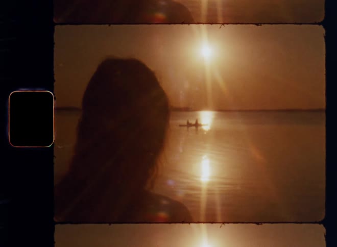 A young woman is standing on the shore watching the sunset over the water with an orange sky as two people are rowing a boat on the lake and the sun creates a bright lens flare on vintage 8mm film with a nostalgic vibe