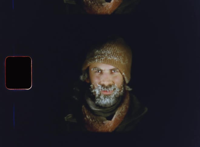 A motivated young man with frozen beard and eyebrows wearing a beige hat covered in snow due to freezing temperatures in a dark winter night looks into the camera in a determined and happy way representing perseverance endurance and stamina