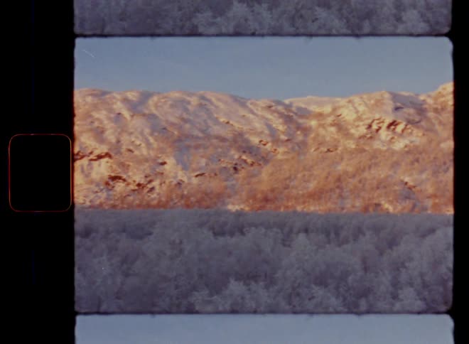 Super 8mm panning wide shot of stunning view of a mountain ridge in a snowy countryside during the day as the winter sun sets and makes the hillside glow in vibrant orange and red colors as the forest remains in the shadow providing contrast