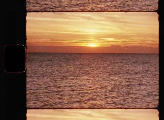 Wide angle shot of open sea at dusk with light waves and the setting or rising sun mostly obscured by horizon during sunrise or sunset on vintage film footage