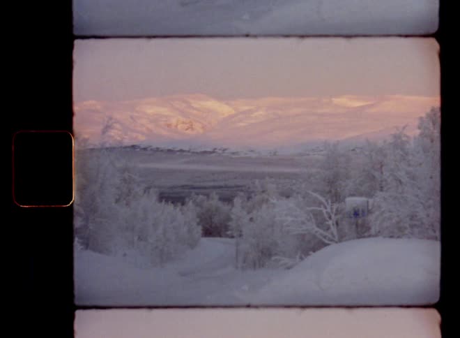 A frozen road leads into a snowy valley landscape covered in fog or mist on a cold and misty January evening surrounded by mountains in the distance as the sun shines onto the hillside making the slope glow red and orange during a beautiful winter sunset