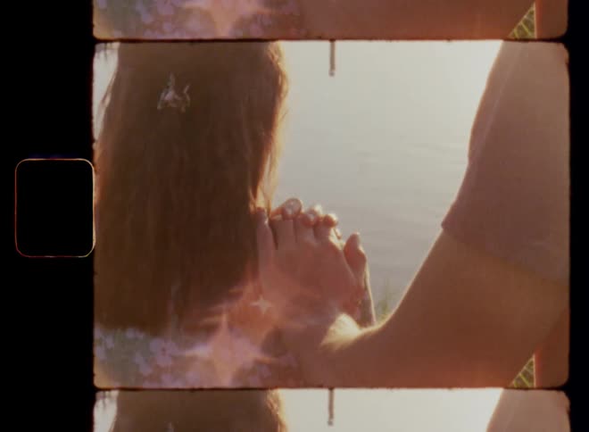 Handheld 8mm film footage of a brunette woman looking out over water as a young man that stands behind her puts his hand on her shoulder and she holds his hand while falling in love