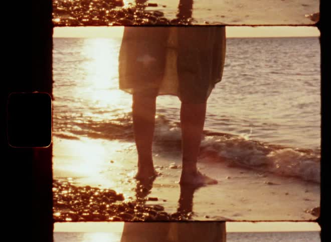 A young woman or girl standing in the water at the beach contemplating and touching the sand and waves with her bare feet as the bright and glaring sun shines and reflects on the water