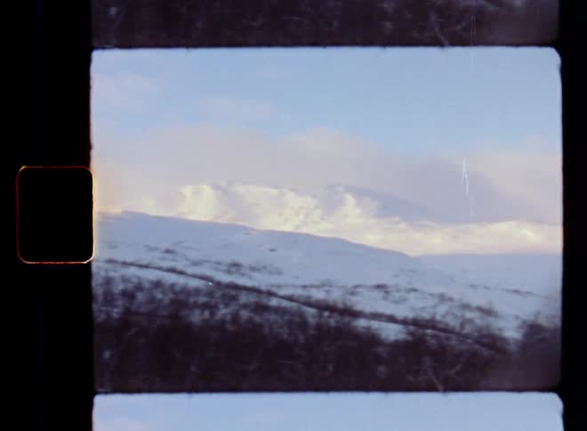 Vintage panning Super-8 film footage of a mountain panorama with a bare forest and leafless trees covered in snow and ice during the icy cold winter above the arctic circle as the low January sun shines onto the wild hills and the valley