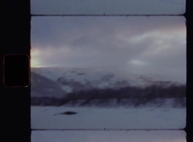 Panning 8mm wide shot or ws of a snowy mountain region with bare trees and snow-covered hill tops or peaks and a white snowy valley with frozen river or lake as dark clouds are in front of the blue sky in the last daylight