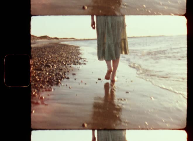 A barefoot woman wearing a light transparent summer dress or skirt walks through the tide on a beach during the day as the sun shines and the waves make her feet touch the warm water of the sea