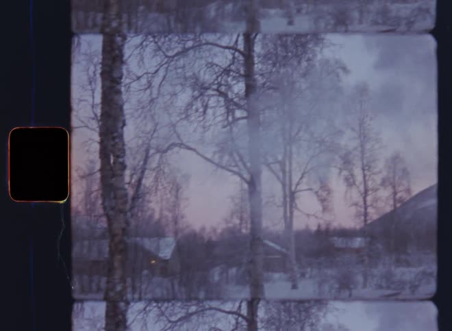 Smoke comes out of a chimney on snow covered roof of cosy fireplace inside small wooden log cabin or holiday home as the wind blows the fume toward the snowy yard with leafless trees in the neighbourhood of mountain village with multiple houses