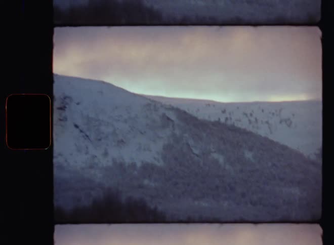 The setting sun shines onto the glowing clouds behind a high mountain ridge or range covered in snow with bare forest in pristine nature at dusk with upward diagonal camera movement shot on vintage Super 8mm camera