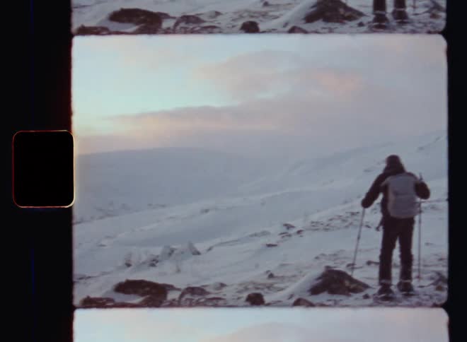 Old super 8mm film footage of a man hiking across a snowy and rocky region of a mountain peak or range during a scientific arctic expedition or field trip on a freezing cold winter day in pristine and untouched nature covered in ice and snow