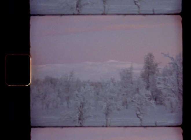 A vintage Super-8 pan shot of snow covered land and trees on a mountain plateau with high altitude against a pink dusk sky as the low winter sun sets and the cold temperatures sink well below freezing