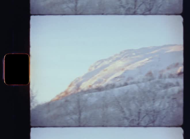 Viewing a white snowy mountain peak through the frozen bare trees of a birch forest on a cold winter day as the sun shines onto the slope of the hill with snow-covered cliffs and rocks under a clear sky in freezing cold temperatures on S8 film footage