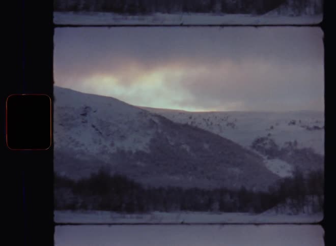 Dark clouds and blue evening sky on a freezing cold overcast winter day with snow storms over a snow-covered valley with frozen trees without leaves surrounded by snowy mountains with high peaks and summits on 8mm retro film