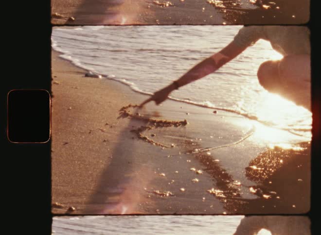 A young unrecognisable woman wearing a light summer dress kneeling at the edge of the sea drawing a heart in the sand of the beach as the waves wash and roll up the shore on old film footage