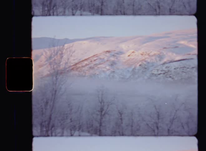 Panning 8mm retro film footage of snow covered mountain range with iced up trees as the mist or fog settles in the valley while the warm morning sun shines onto the peaks which glow in the sunrays during beautiful winter sunrise