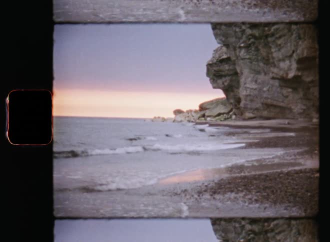 An ocean coastline on a gloomy day with clouds and beautiful sunset with waves rolling onto the beach in front of a tall rock face or steep cliff formation at the high coast