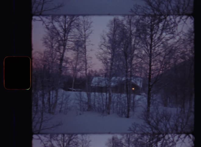 A holiday village consisting of small cottages or cabins in a bare forest in a mountain valley during sunset in last daylight at dusk on a cold winter day with snow and ice and snow-covered roofs on vintage Super-8 film footage