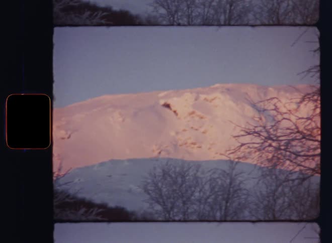 Panning Super 8mm shot of a white snow covered south face of a mountain ridge on the countryside during the day as the sun sets creating a beautiful contrast between the shadow in the valley and the bright and glowing hillside