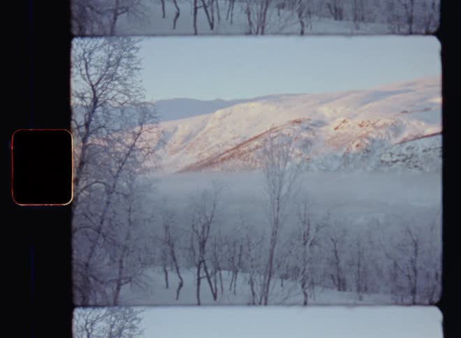 Static handheld 8mm film footage of snowy mountain range or peaks with snow covered trees as the sun sets and the fog or mist rolls into the valley with wild and untouched pristine nature and real winters back in the days with temperatures below freezing