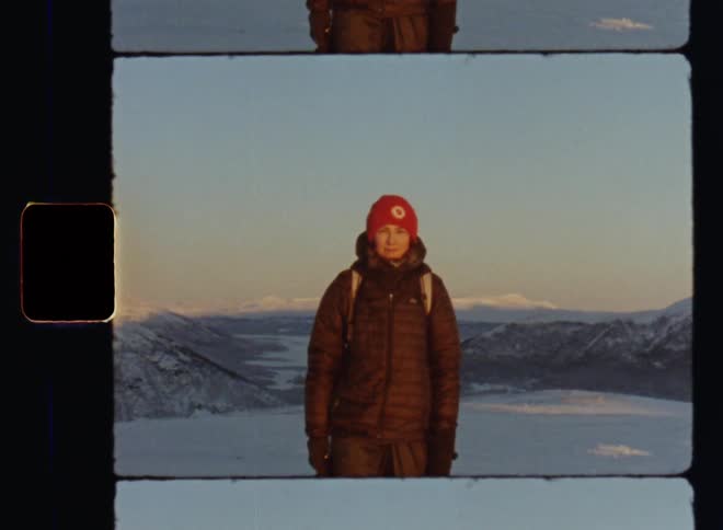 Young brunette woman is smiling and standing on a hill top or peak wearing winter hiking gear with mountains and valleys covered by snow in the background as the rising morning sun warms her happy face on Super-8 mm film