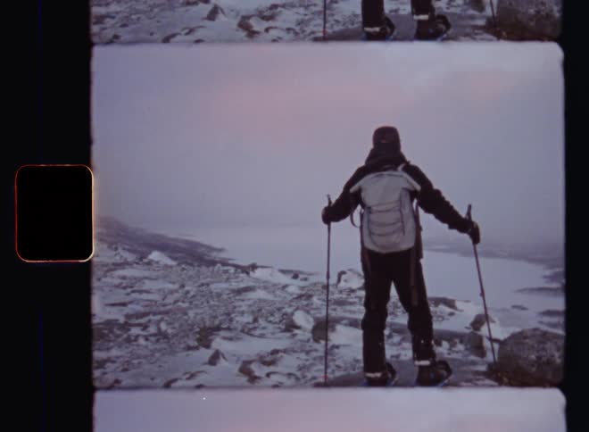A man with backpack snow shoes and ski poles or sticks stands on the edge of a cliff enjoying the view over the valley and mountains in winter wonderland as a blizzard or snow storm with dark clouds rolls in on a cold December or January evening up north