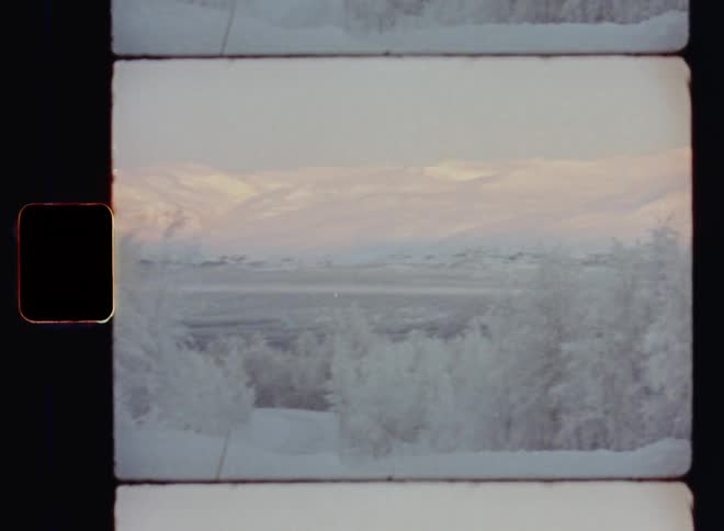 Panning shot with horizontal movement of a snowy winter wonderland landscape with iced up and snow-covered trees as the sun shines onto distant mountain peaks in the arctic north filmed on Super 8mm with vintage film burn