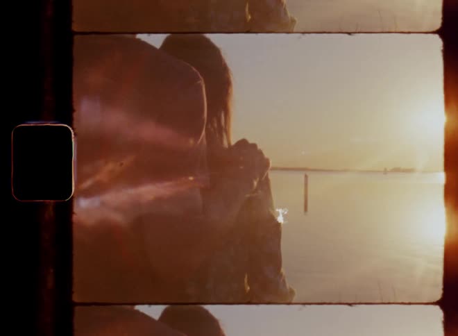 A man lays his hand on the shoulder of a young woman with brown hair while standing together in front of the water watching the sunset during a romantic honeymoon date on nostalgic 8mm film with lens flare