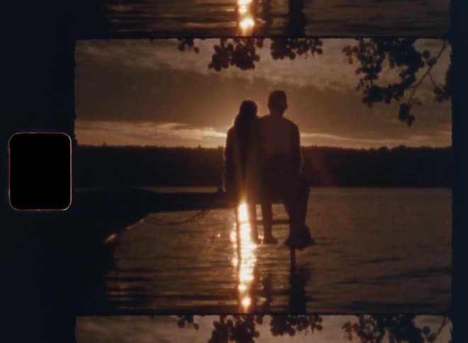 Super 8mm Ws of the silhouette of a romantic couple sitting on the edge of a pier next to a boat at the shore of a forest lake surrounded by trees at sunset with back light and bright lens flare